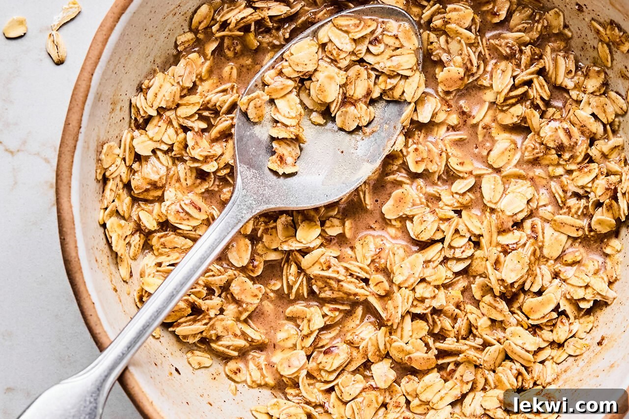 A bowl filled with the mixture of oats, protein powder, cinnamon, maple syrup, almond milk, and coconut oil after being stirred with a metal spoon.