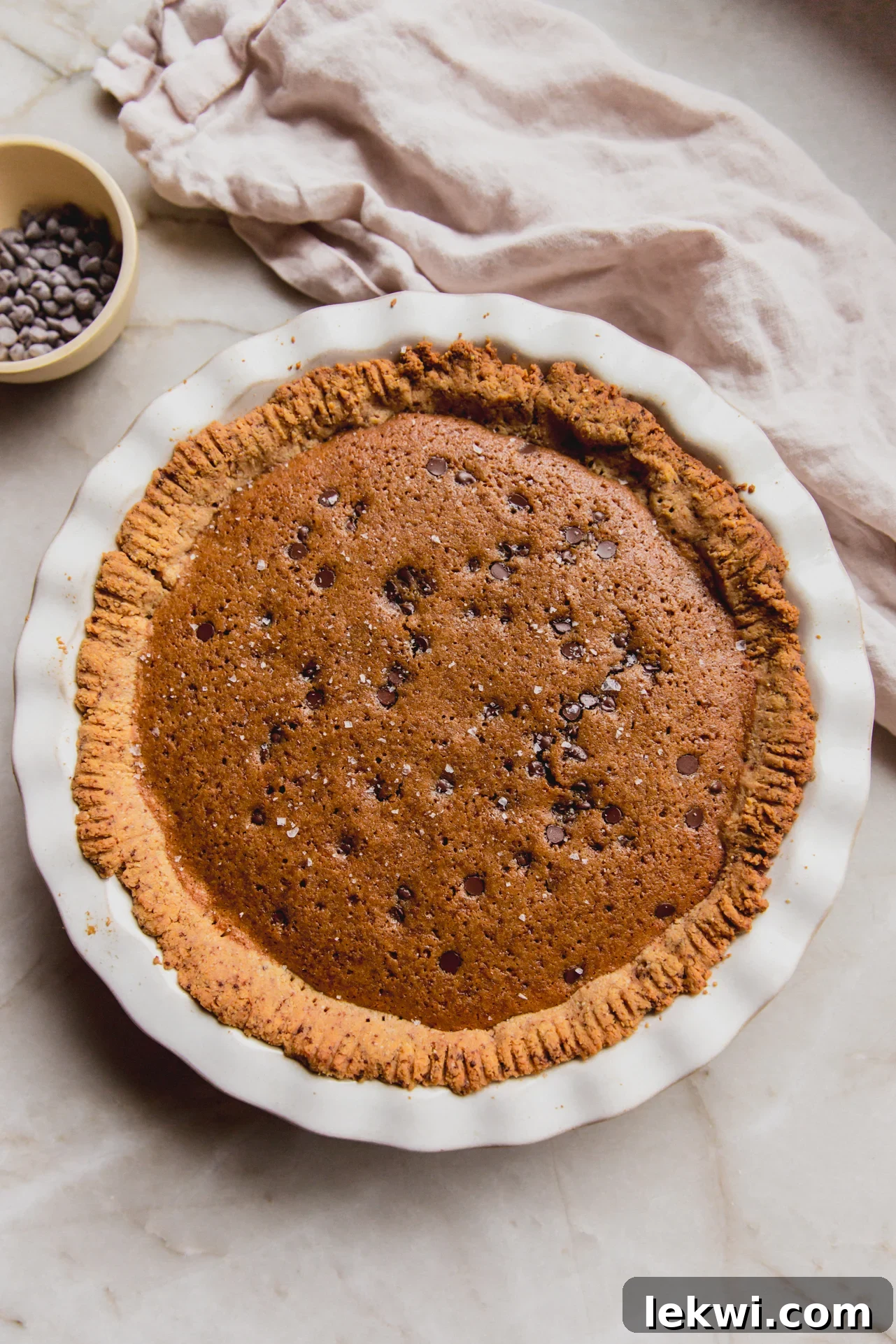A perfectly baked gluten-free chocolate chip cookie pie in a white ceramic pie pan, with a small bowl of chocolate chips beside it, ready to be served.
