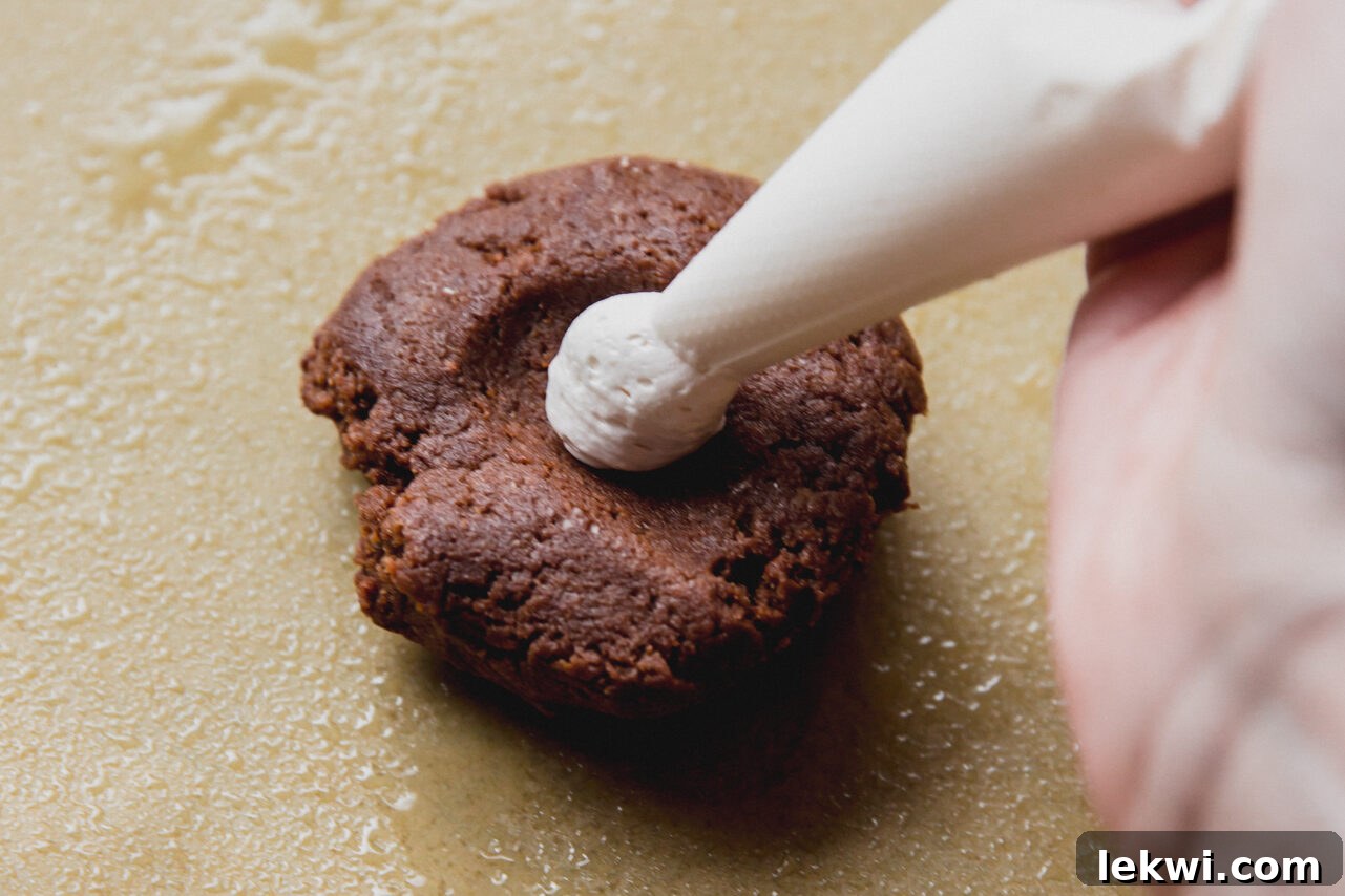 A hand piping marshmallow fluff directly into the thumbprint indentation of a chocolate cookie dough ball on parchment paper.