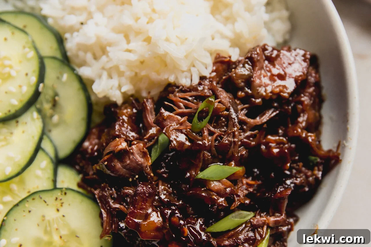 A white bowl with ginger chuck roast next to white rice and cucumber slices.