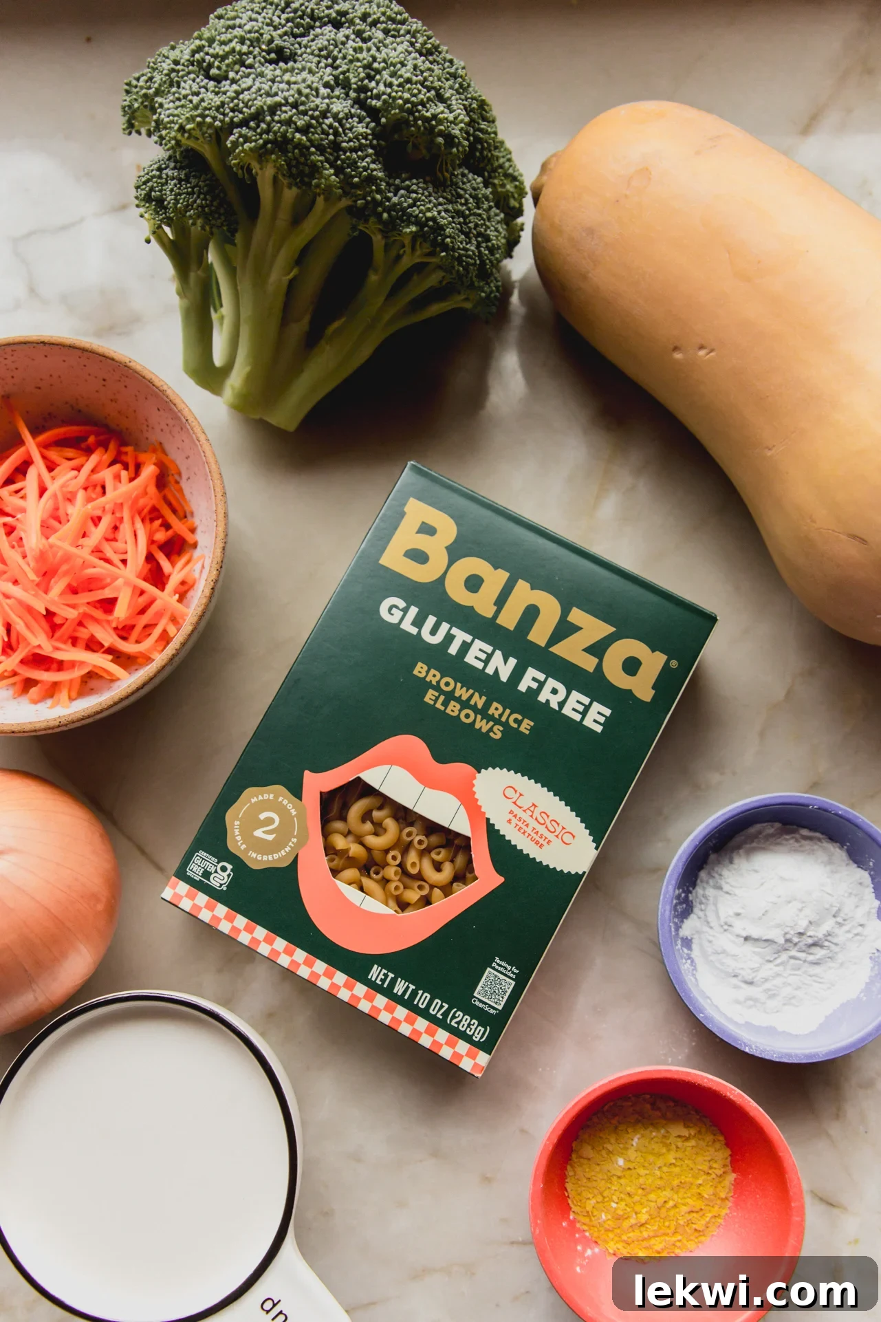 Fresh ingredients for broccoli cheese noodle soup, including broccoli florets, carrots, onions, garlic, butternut squash, and Banza pasta, are neatly arranged on a clean marble countertop.
