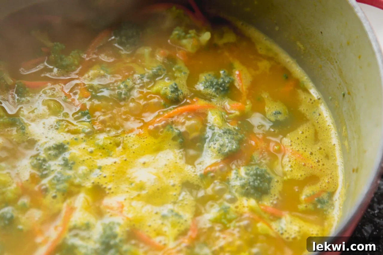 Broccoli cheese soup simmering in a large pot, with vibrant green broccoli florets and bright orange shredded carrots, adding color and texture.