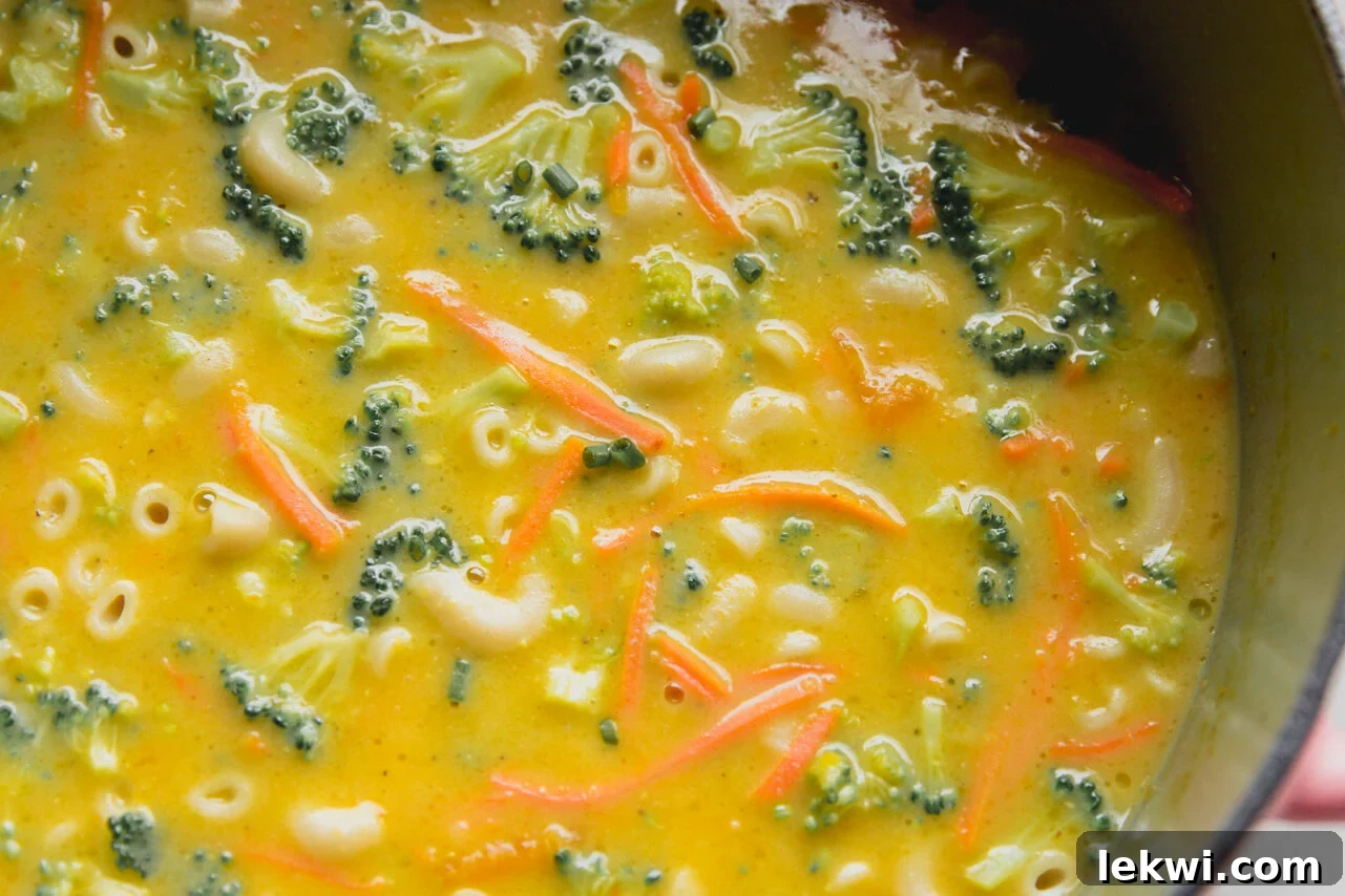 Creamy broccoli cheese noodle soup, rich with broccoli florets, shredded carrots, and gluten-free noodles, stirred in a large pot, ready to be served.