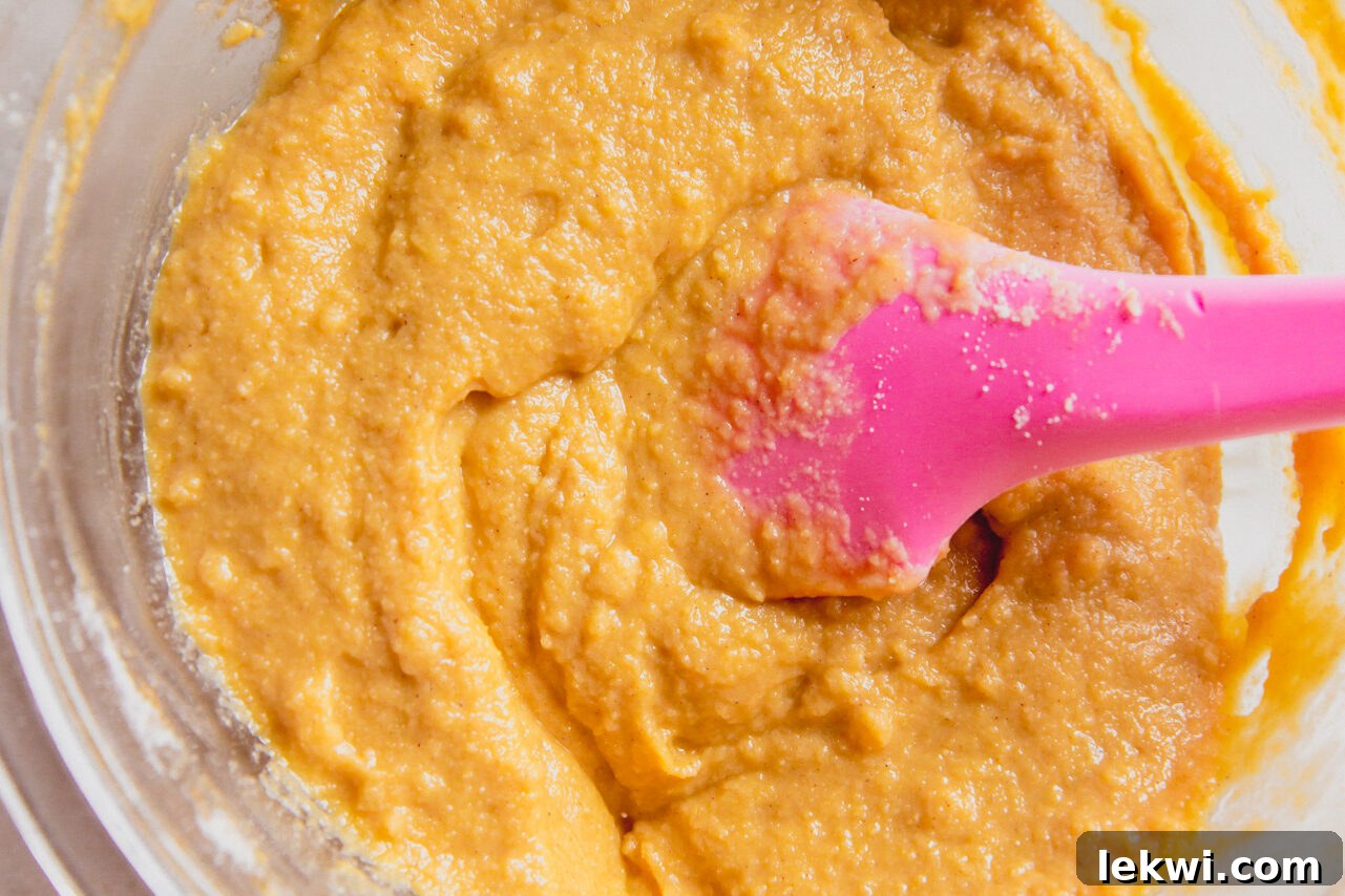 A glass bowl filled with a light, creamy cinnamon roll cake batter, with a pink spatula resting in it.