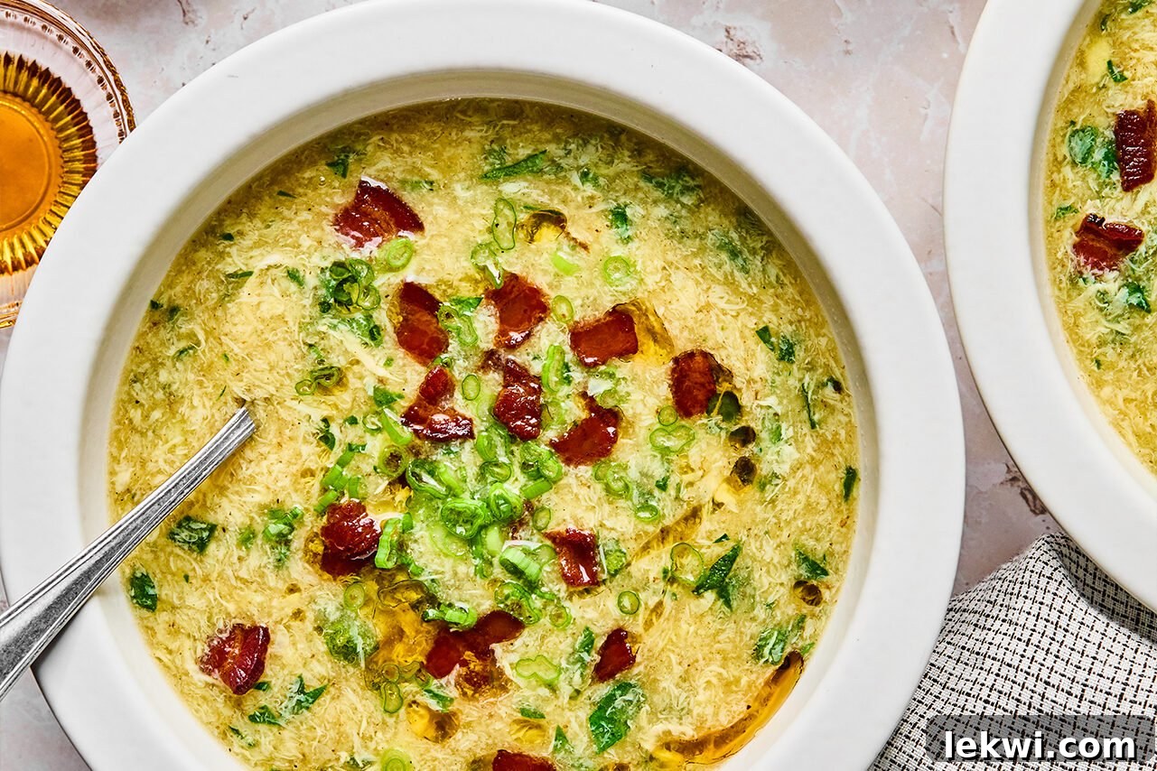 A beautifully presented bowl of Breakfast Egg Drop Soup, generously topped with crispy bacon bits, a drizzle of sesame oil, and freshly chopped green onions, with additional bacon on the side.