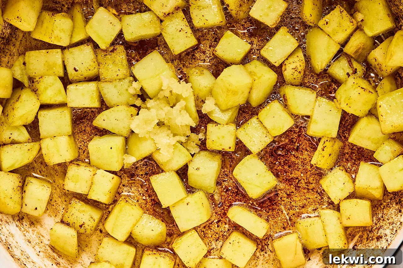 Diced sweet potato actively sautéing in a pan, with minced garlic freshly added on top, contributing to the fragrant base of the soup.