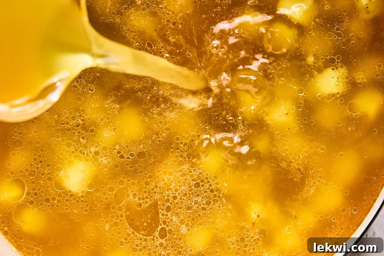 Chicken bone broth is poured into a pot containing sautéed sweet potatoes, as a slurry of arrowroot starch mixed with water is slowly streamed in, being constantly stirred to thicken the broth.