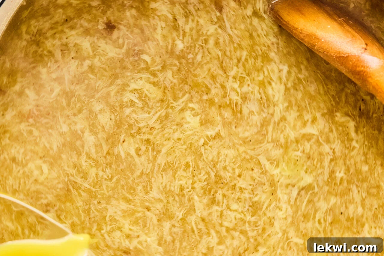 A Dutch oven filled with simmering soup, showing delicate egg ribbons forming as whisked egg is slowly streamed in while stirring with a wooden spoon.