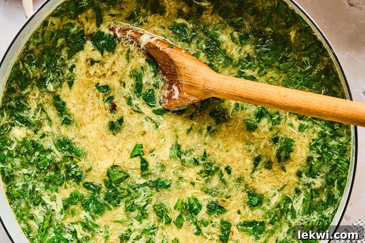 The finished soup in a Dutch oven, with fresh spinach just added and wilting into the hot broth, being stirred gently with a wooden spoon.