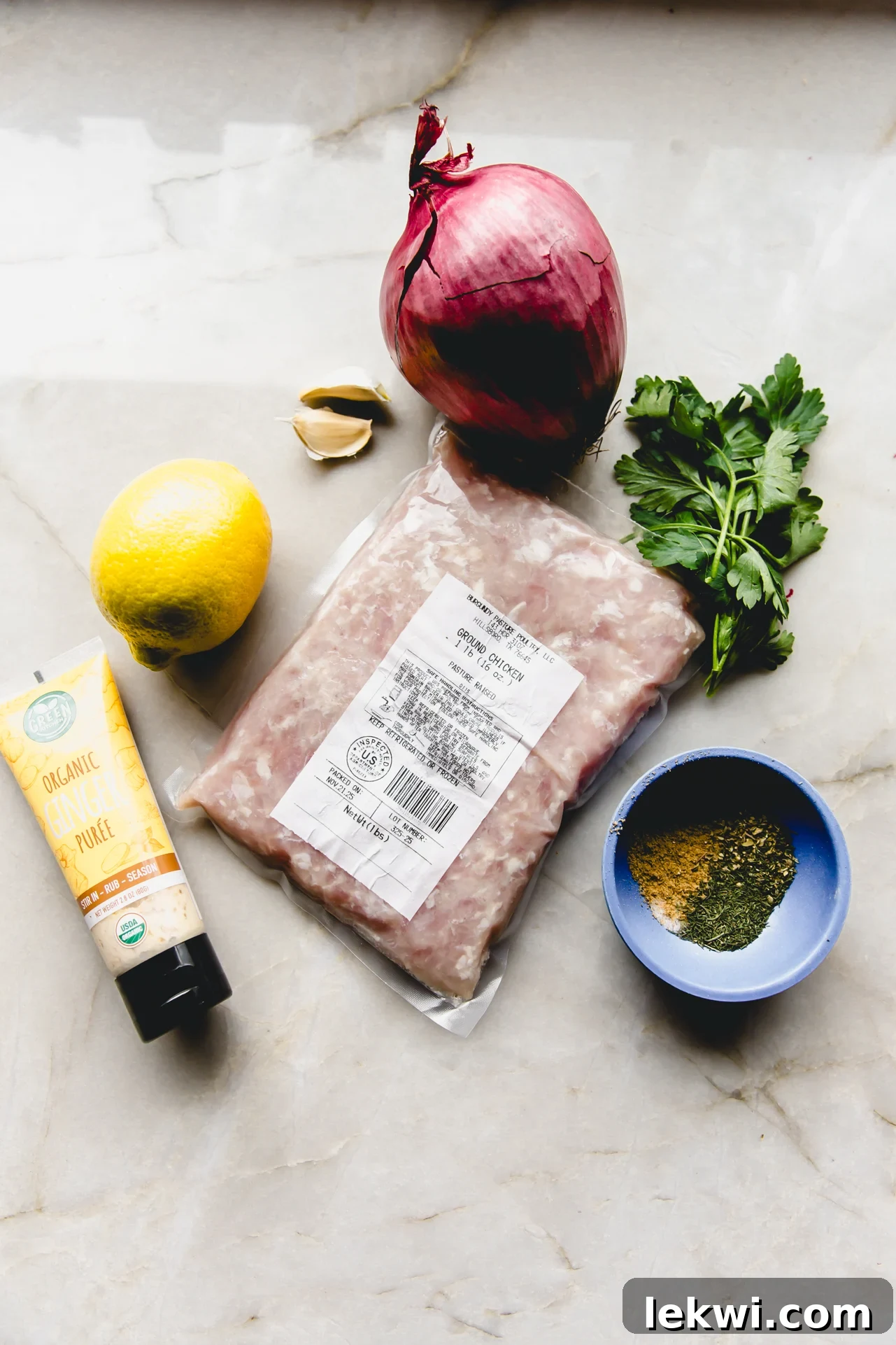 Sheet pan kebab ingredients laid out on a marble counter top, including ground chicken, spices, red onion, garlic, and lemon.