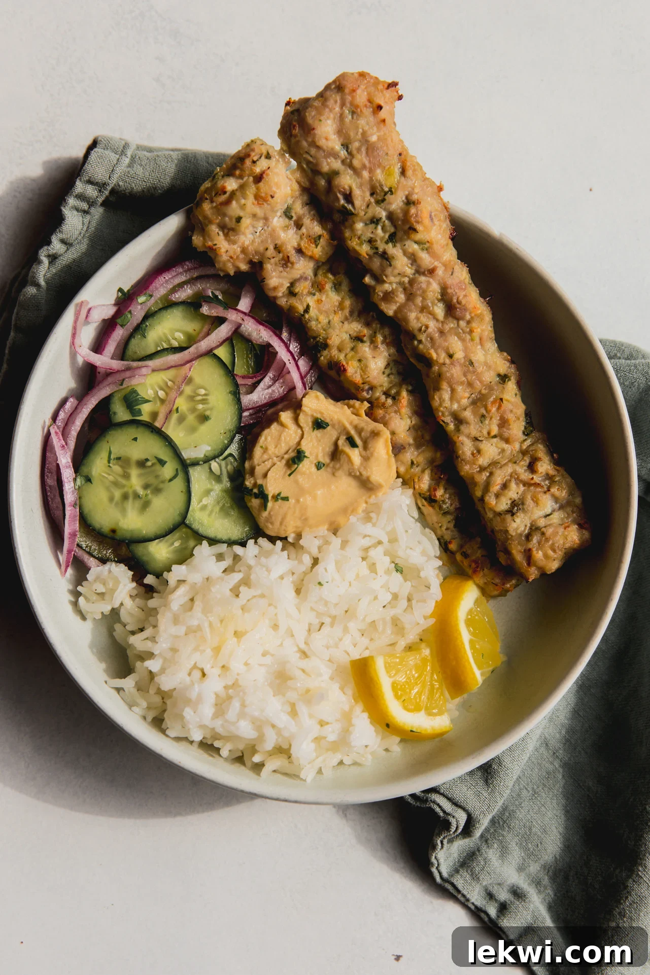 Two sheet pan kebabs in a bowl with cucumber salad, white rice and sliced lemon wedges.
