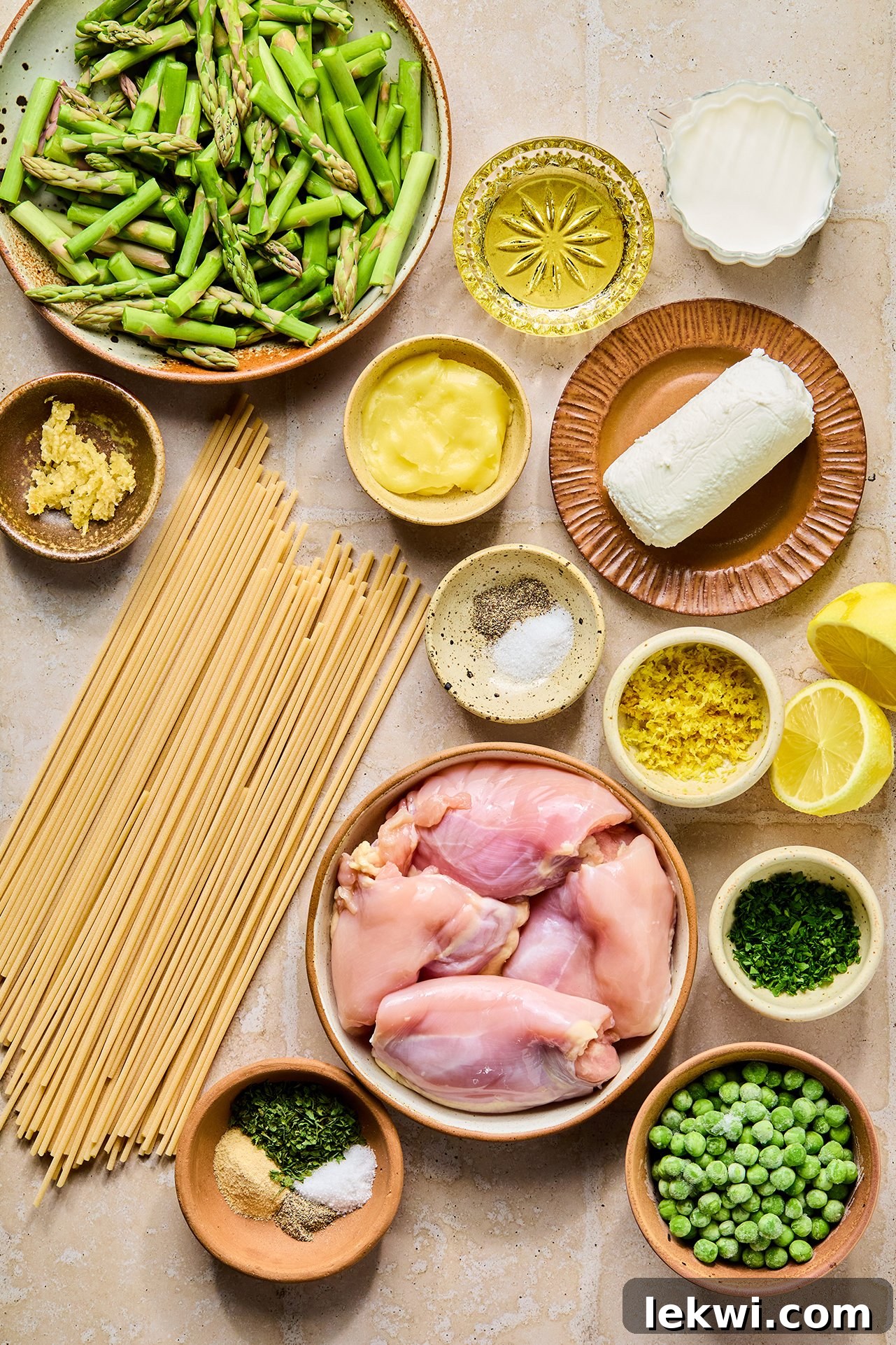 All the fresh ingredients for lemon goat cheese pasta, including chicken, asparagus, peas, lemon, garlic, and goat cheese, laid out in bowls on a kitchen counter before cooking.