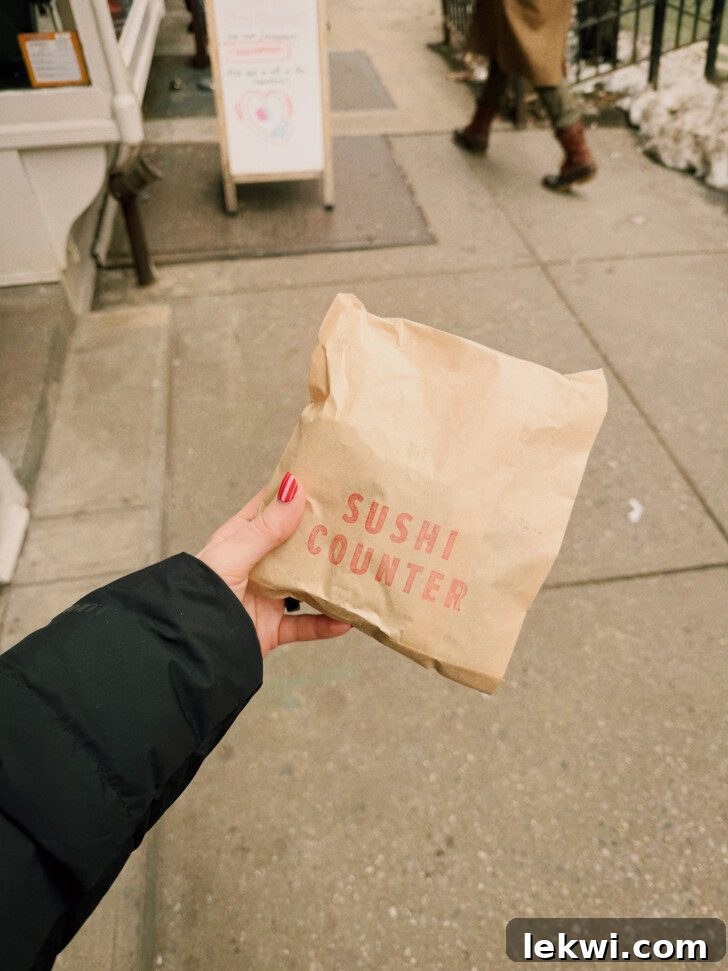 A hand holds a brown paper bag from "Sushi Counter" against a blurred city sidewalk background, suggesting a grab-and-go meal in an urban setting.