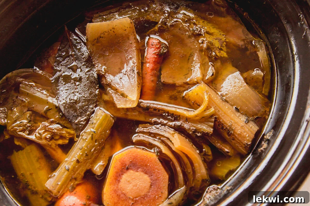 A slow cooker filled with all the bone broth ingredients: roasted beef bones, colorful vegetable scraps, aromatic fresh herbs, and water, poised for a long, nourishing simmer.