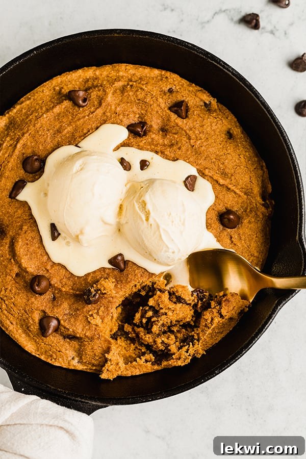 Spoon scooping a warm, gooey pumpkin chocolate chip cookie from a mini cast iron skillet, topped with melted ice cream.