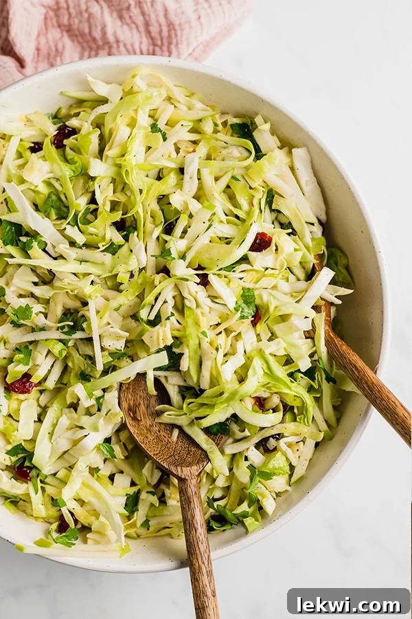 A large white serving bowl filled with vibrant Thanksgiving coleslaw, ready to be served, with two wooden spoons for dishing.