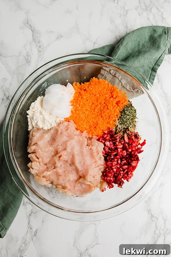 A large mixing bowl containing all the raw ingredients for the turkey poppers: ground turkey, shredded sweet potato, chopped cranberries, and a vibrant medley of fresh rosemary, sage, and parsley.