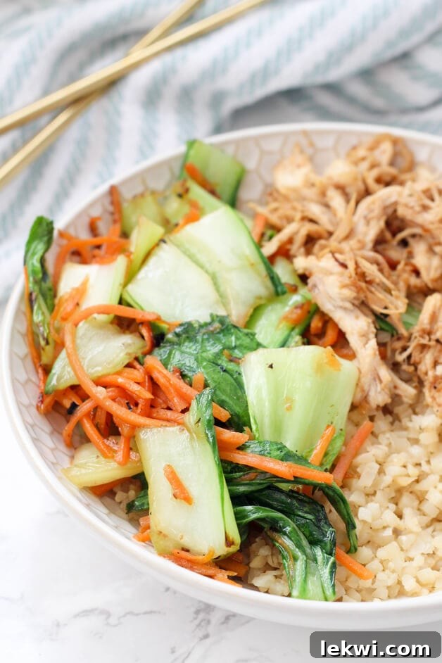 Simple Paleo, Whole30, and AIP Chicken and Vegetable Stir-Fry 3 A close-up shot of a white bowl filled with a colorful chicken and vegetable stir fry, featuring shredded chicken, bright orange carrots, green baby bok choy, and fluffy cauliflower rice.