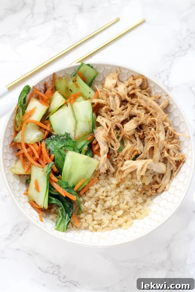 A vibrant and colorful chicken and vegetable stir fry dish presented in a modern white bowl, ready to be enjoyed. The dish features shredded chicken, cauliflower rice, baby bok choy, and shredded carrots, all coated in a savory sauce.