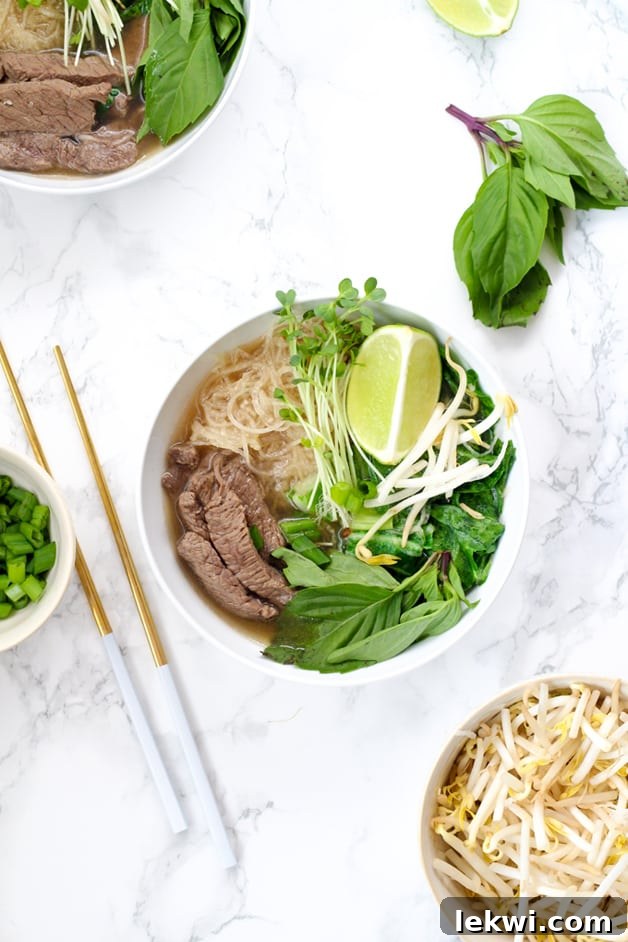 A bowl filled with pho including beef, rice noodles, herbs, bean sprouts, limes, and chop sticks next to it.
