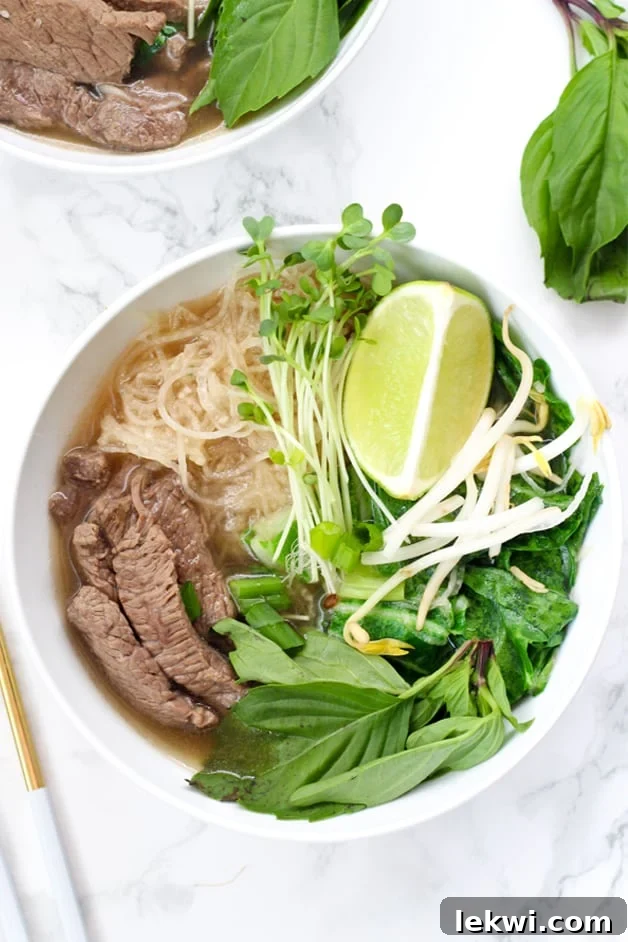 A white bowl filled with beef pho with rice noodles, herbs, lime, and meat.