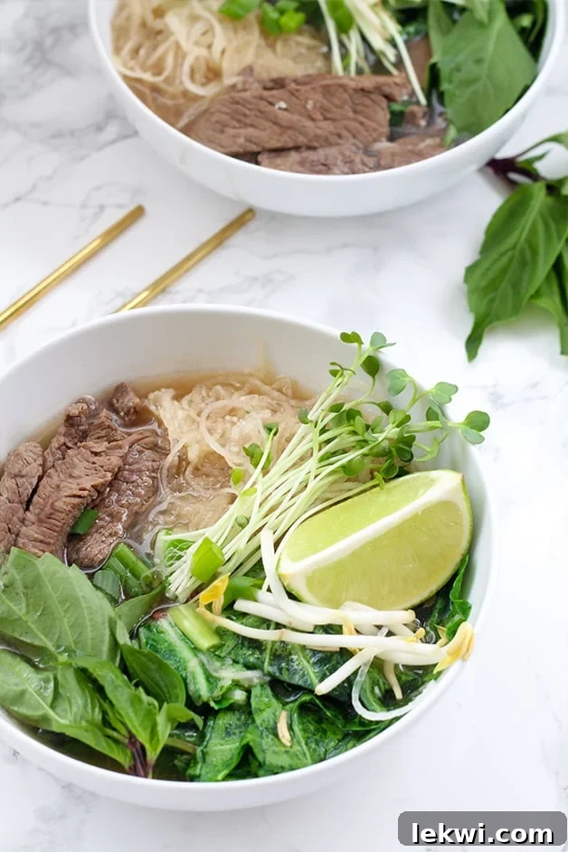 A bowl filled with pho including beef, rice noodles, herbs, bean sprouts, limes, and chop sticks next to it.