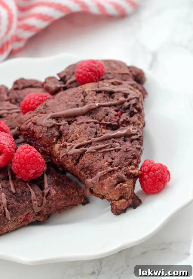 A white plate displaying three homemade Paleo Chocolate Raspberry Scones, garnished with fresh raspberries and a light dusting of cocoa powder.