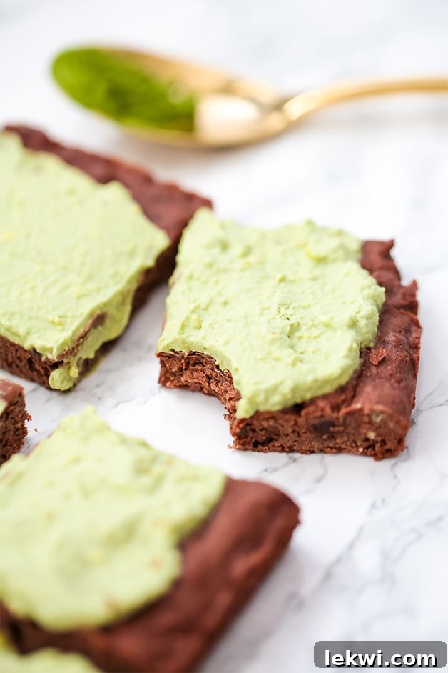 A few AIP Paleo Matcha Brownies with bright green frosting displayed on a rustic counter, one with a delicious bite taken out.