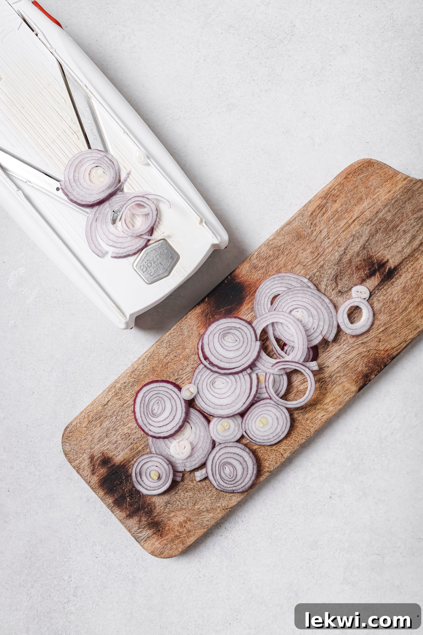 A mandoline slicer being used to thinly slice a red onion on a cutting board.
