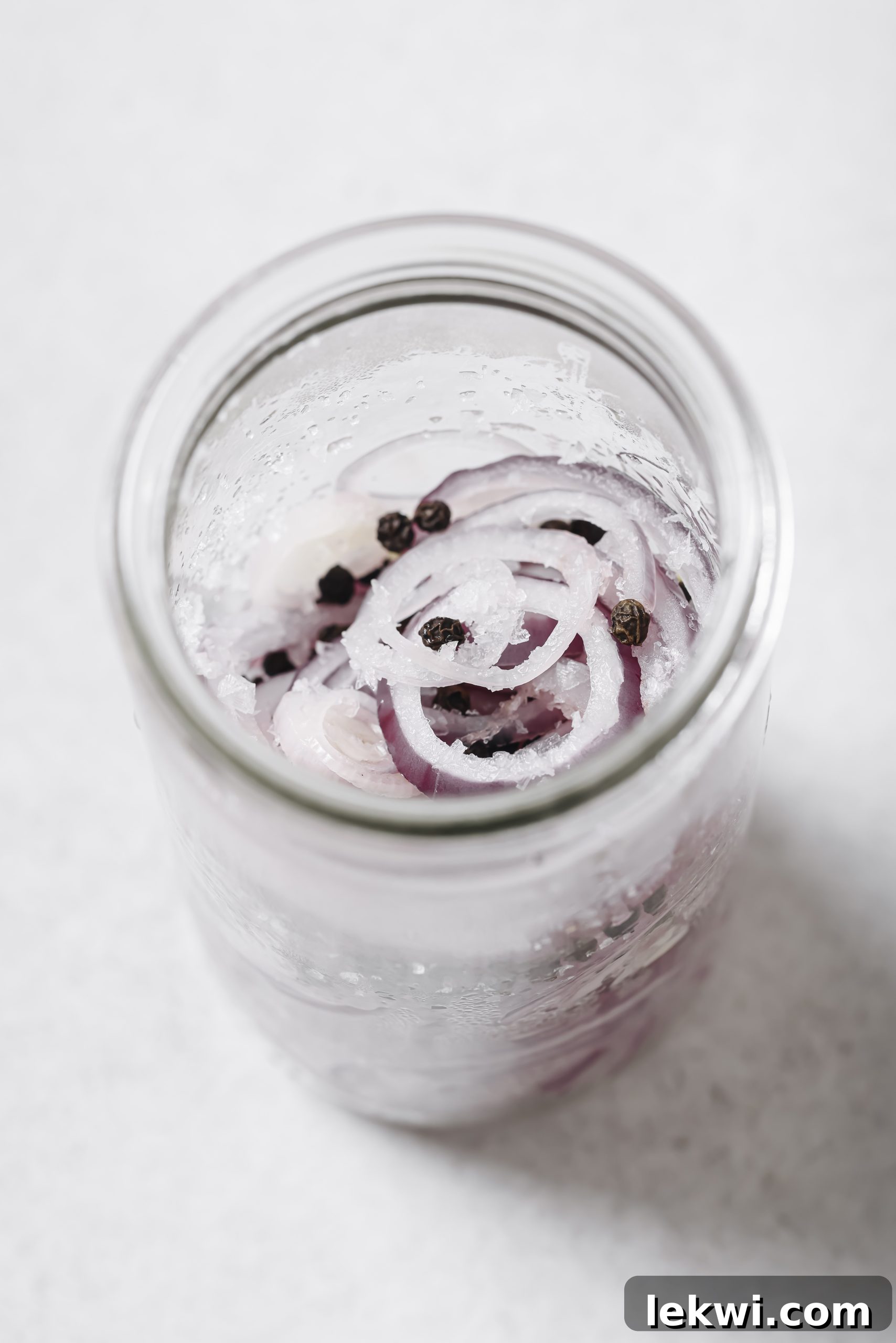 Red onion slices being placed into a jar with peppercorns and salt.