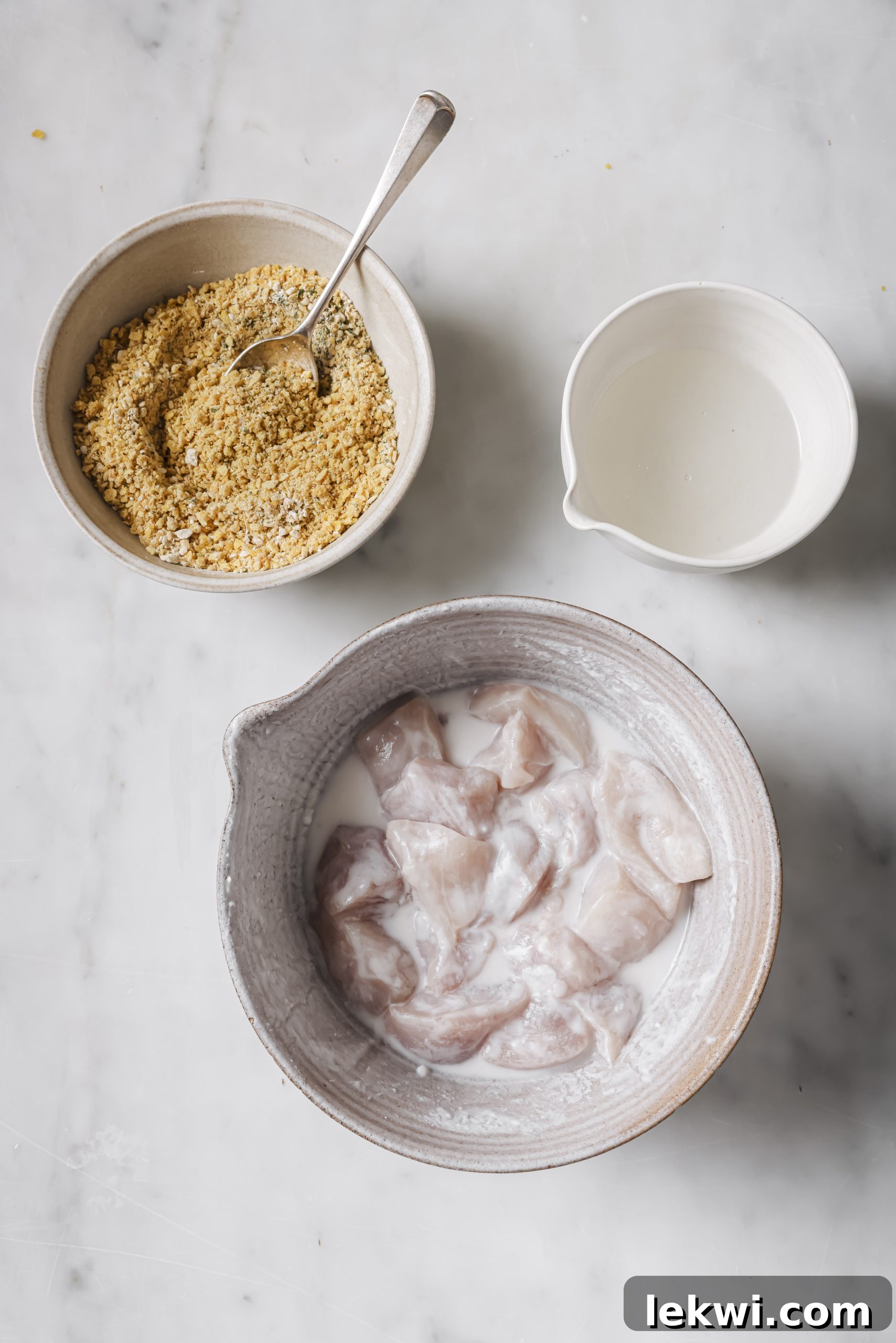 Three separate bowls arranged as a dredging station: marinated chicken, melted coconut oil, and seasoned plantain chip crumbs.