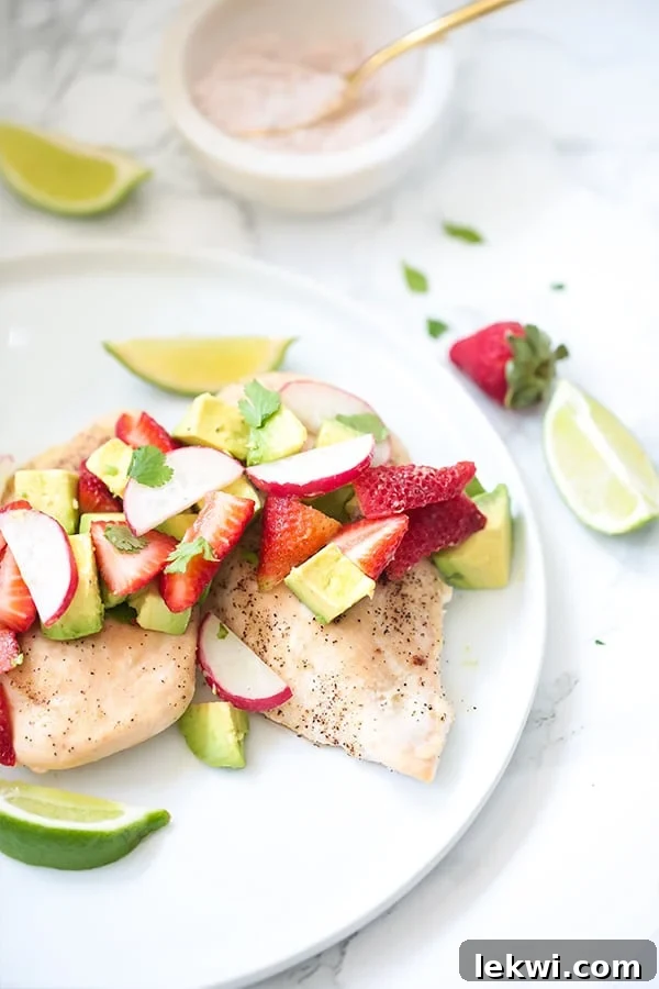 A white plate with cooked chicken breast covered in strawberry and avocado with lime wedges next to it.