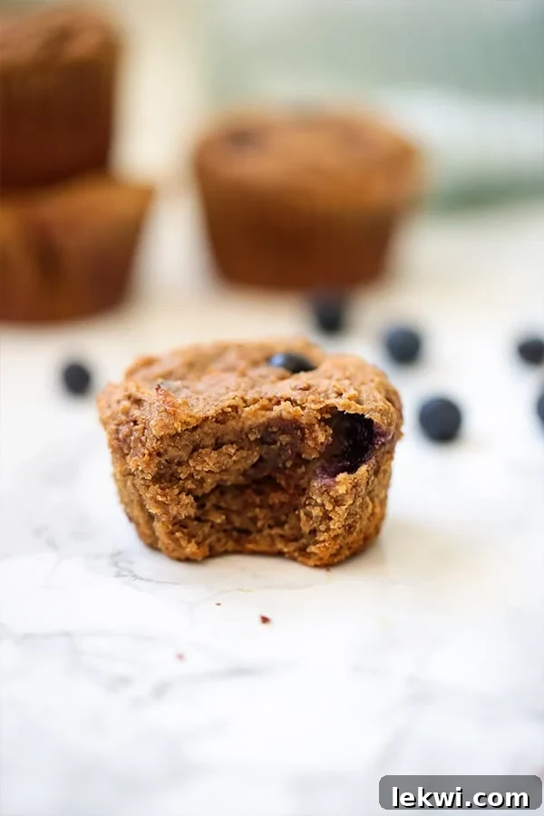 A close-up of a single blueberry banana muffin, with a bite taken out, revealing its fluffy interior, alongside scattered fresh blueberries. It emphasizes the appealing texture and fresh ingredients.