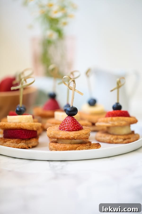 A white plate featuring a close-up of colorful mini pancake skewers interspersed with fresh berries and banana slices