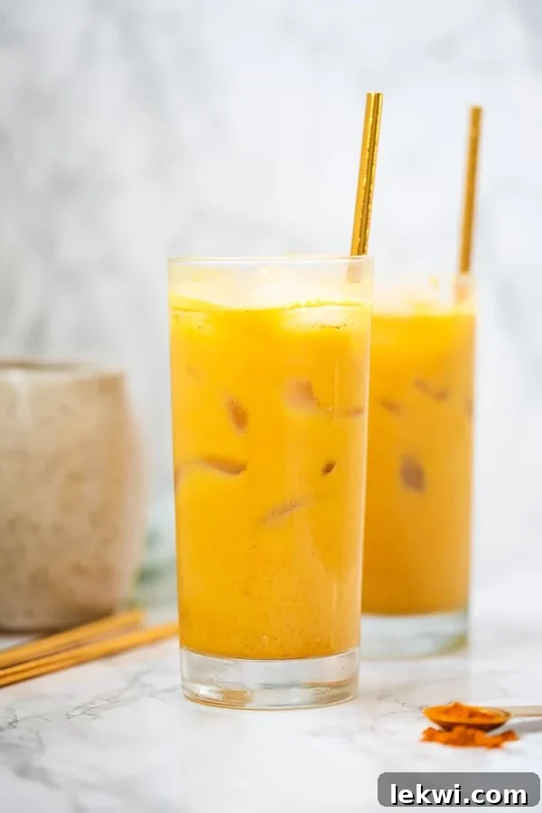 Two glasses filled with iced turmeric golden milk lattes, adorned with fresh mint and straws, resting on a rustic wooden surface with scattered turmeric powder and cinnamon sticks.
