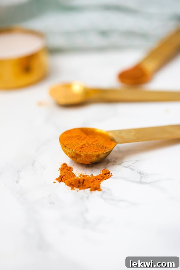 Close-up of a vibrant golden turmeric root, partially sliced, surrounded by whole turmeric roots, spices, and a glass of the finished latte in the background.