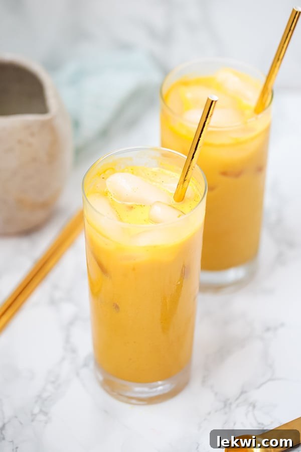 Two hands holding glasses of Iced Golden Milk Turmeric Latte, with a pitcher of ingredients and fresh spices in the background.