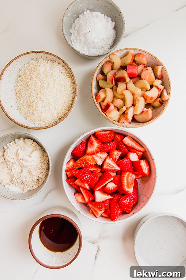 A vibrant display of fresh ingredients for strawberry rhubarb crisp, including ripe, red strawberries, crimson rhubarb stalks, unsweetened shredded coconut, and tigernut flour, meticulously laid out on a clean wooden surface.