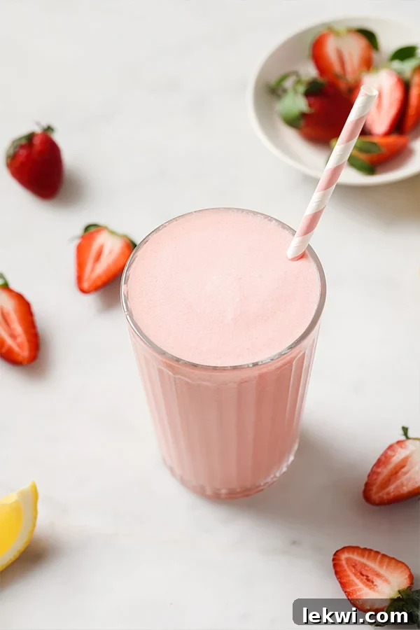 Straw in a tall glass of creamy frozen pink lemonade, garnished with a lemon slice and fresh strawberry.