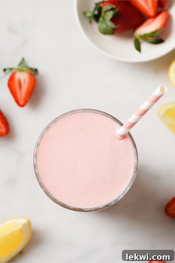 Glass of creamy frozen pink lemonade on a table with fruit, with a wooden spoon and a straw.