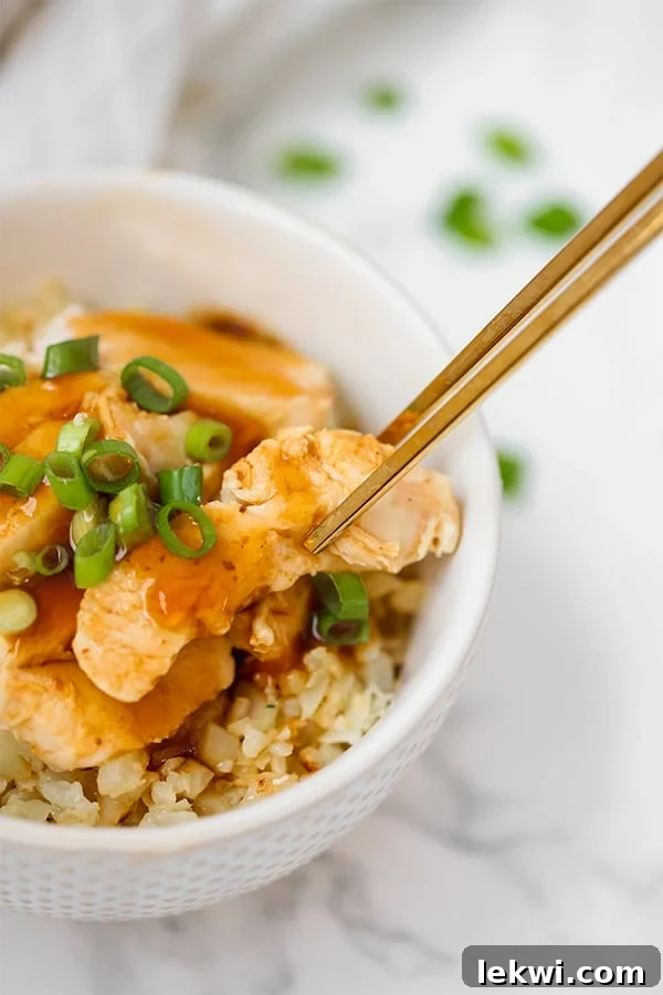 A white bowl with cauliflower rice topped with slow cooker chicken teriyaki, garnished with fresh green onions, with chopsticks ready to pick up a piece.