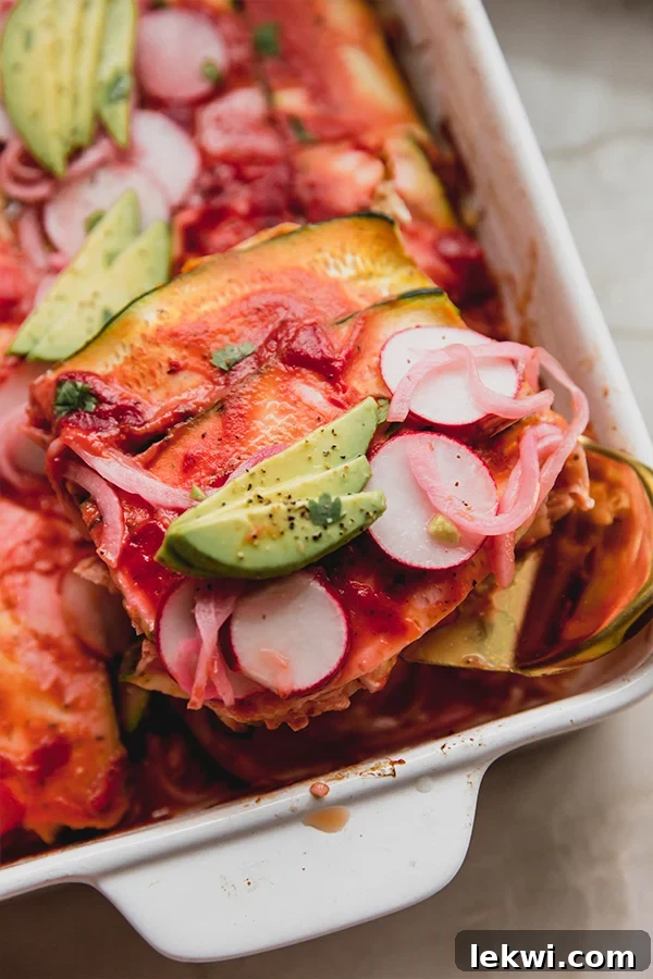 Zucchini enchilada casserole in a baking dish topped off with enchilada sauce, slices radish, avocado, pickled onion and cilantro, with a slice being taken out.