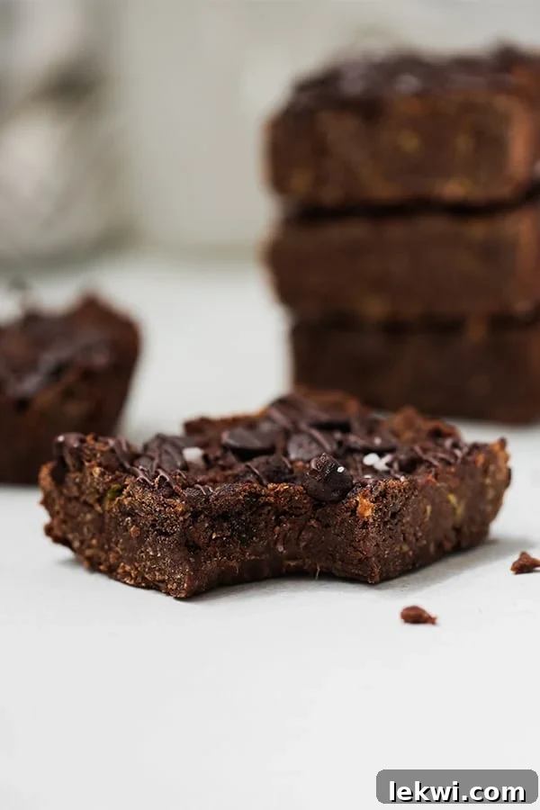 A zucchini brownie with a bite taken out of it in front of a stack of zucchini brownies.