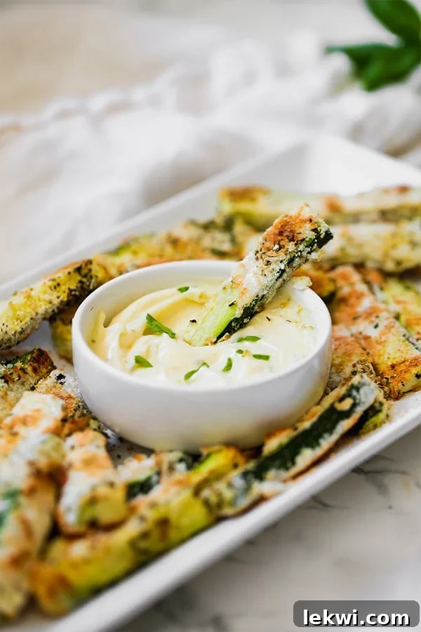 A close-up shot of a single golden-brown baked zucchini fry being dipped into a small white bowl of creamy garlic mayo, surrounded by more fries.