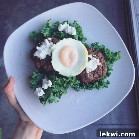 A wholesome dinner featuring two grass-fed beef burgers, a baked egg, and a side of fresh kale with goat cheese.