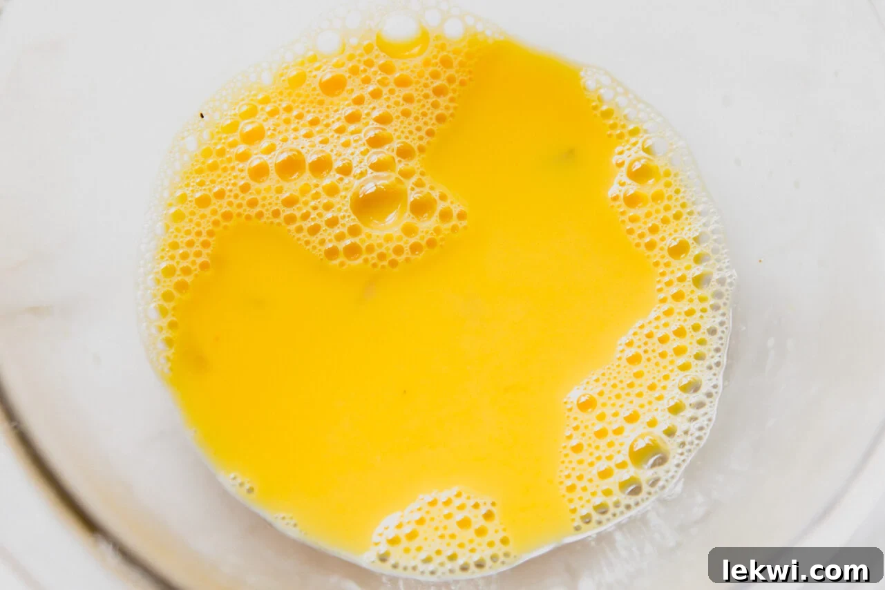 Egg yolks being slowly tempered with hot broth, showing a creamy, emulsified texture forming in a glass bowl.