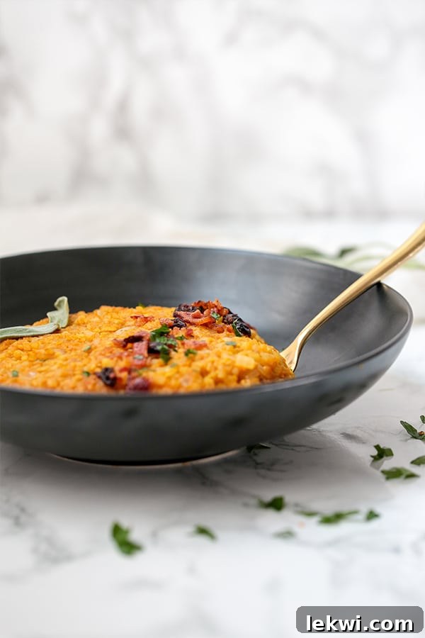 A close-up view of a dark bowl brimming with creamy cauliflower pumpkin risotto, featuring a gleaming metal spoon. The focus is on the rich texture and warm, inviting colors of the dish, hinting at its savory and comforting qualities.