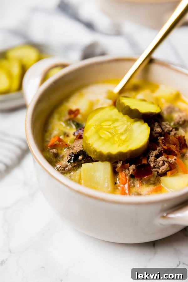 Paleo Bacon Cheeseburger Soup Creamy and Dairy Free 2 A steaming bowl of dairy-free bacon cheeseburger soup, garnished with fresh pickles and a spoon, inviting you to take a bite.