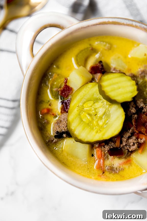 Paleo Bacon Cheeseburger Soup Creamy and Dairy Free 3 A pristine white bowl overflowing with savory bacon cheeseburger soup, adorned with an extra generous serving of dill pickles on top.