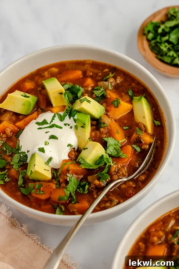 A steaming bowl of paleo pumpkin chili, garnished with fresh herbs, and a spoon ready for a comforting bite, set on a rustic wooden surface.